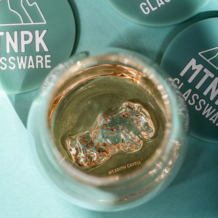 An open round glass jar containing a clear gel or liquid with a metallic rose-gold/copper reflective surface, placed on a teal surface. Partial branded coasters are visible around it.