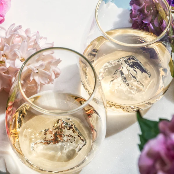 Two glasses of white wine with decorative ice cubes on a white plate, surrounded by flowers.