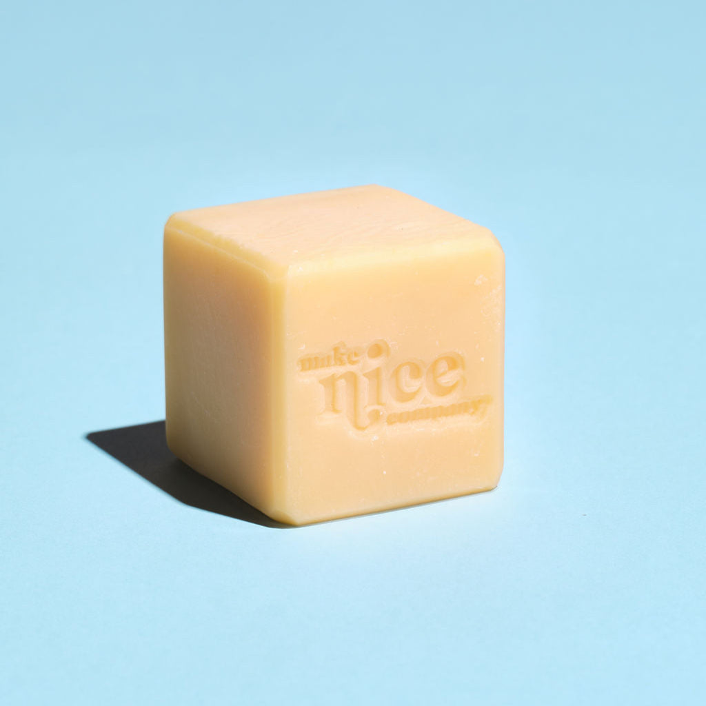 Unwrapped Make Nice Company Citrus Solid Dish Soap bar with an embossed logo, placed on a light blue background. Made with lemon, blood orange, and bergamot.