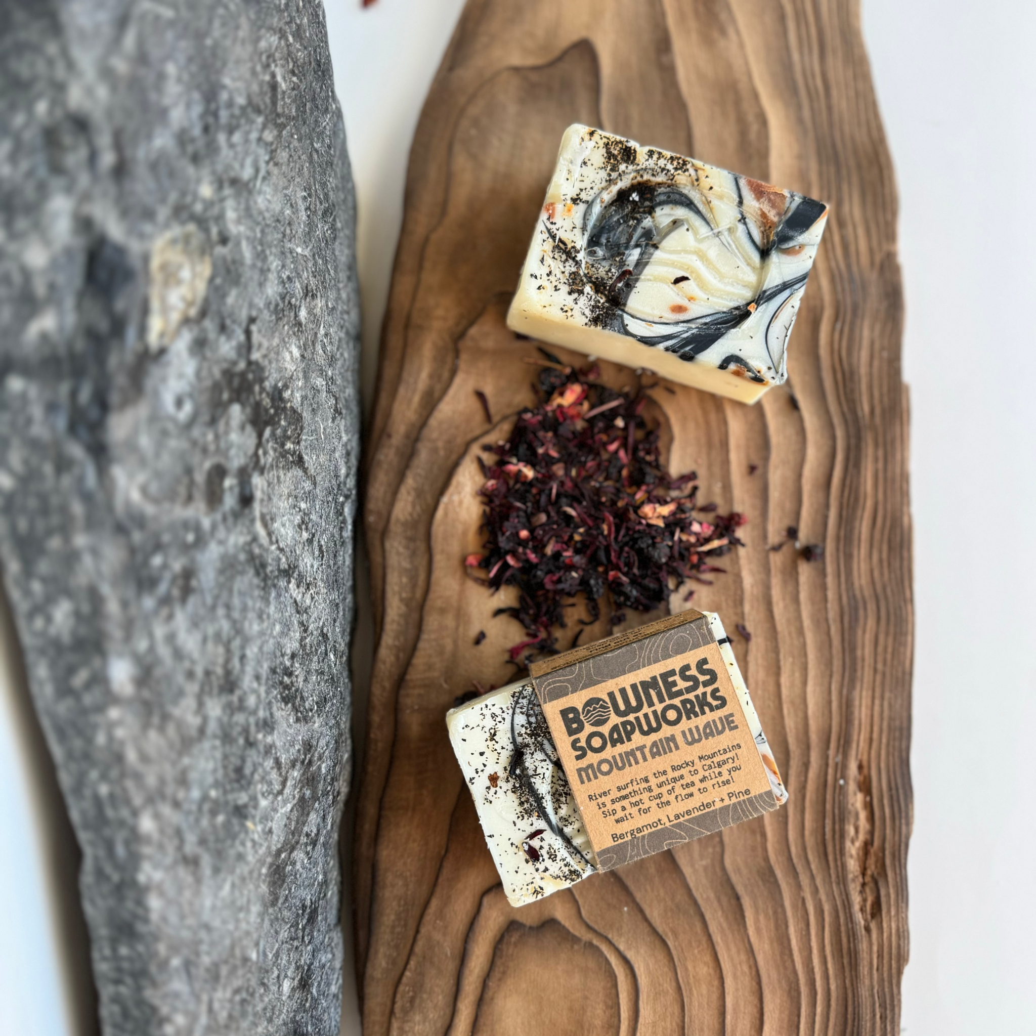 Flat lay of Mountain Wave herbal soap bar by Bowness Soapworks in Calgary, with dried botanicals on a wood board beside a textured rock