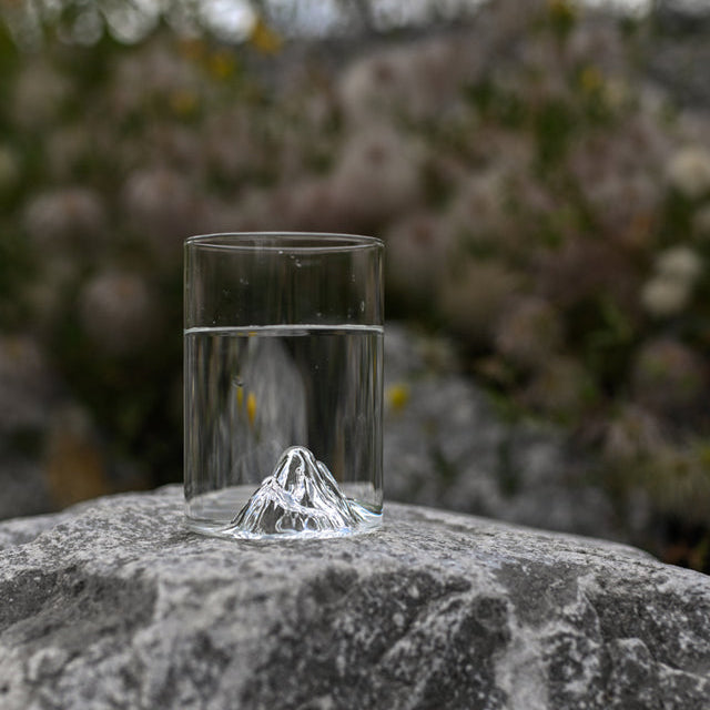 MTNPK pint glass with 3D mountain base resting on grey rock outdoors with blurred foliage in background