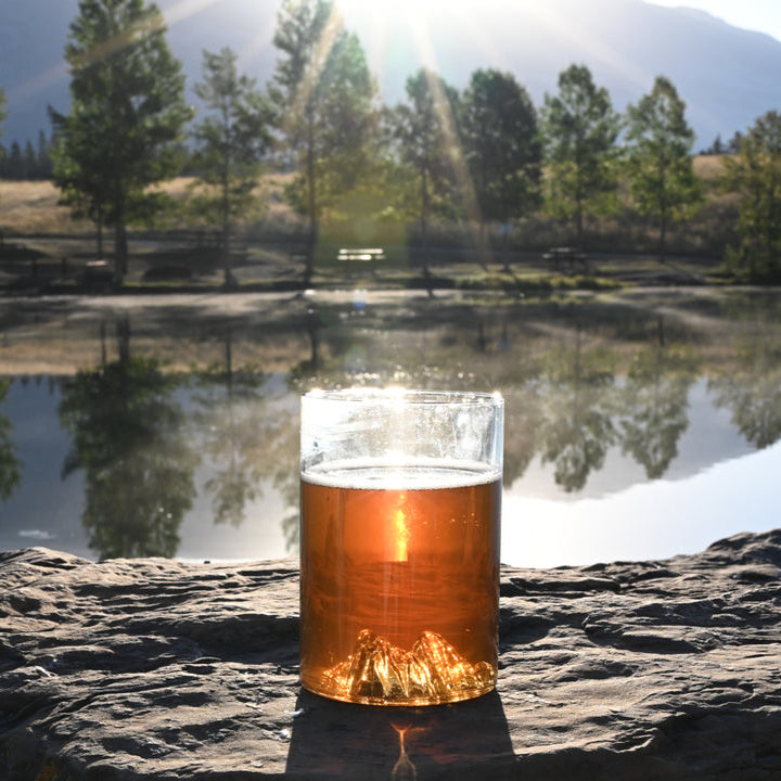 MTNPK Squamish pint glass with amber drink on rock ledge by a lake, sold at Greenbriar Market in Calgary