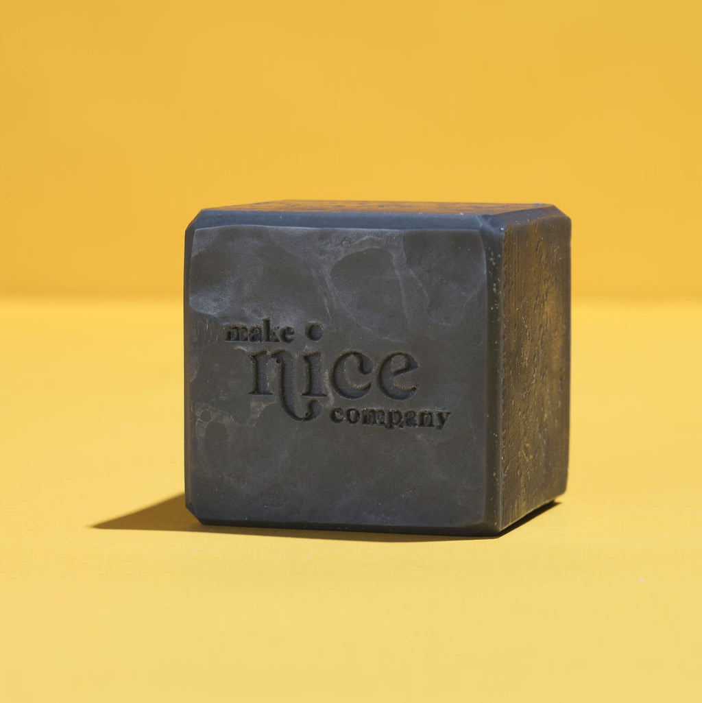 Unwrapped Make Nice Company Charcoal Solid Dish Soap bar, embossed with the brand logo, placed on a yellow background. Made with activated charcoal for deep cleaning.