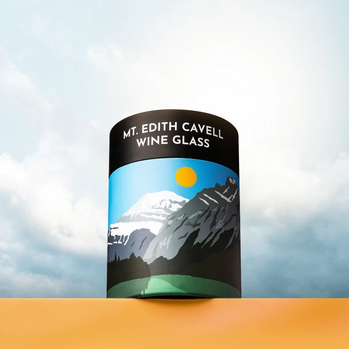 A cylindrical wine glass with a black top band reading 'MT. EDITH CAVELL WINE GLASS' and a mountain landscape label, shown standing on an orange surface against a cloudy sky background.